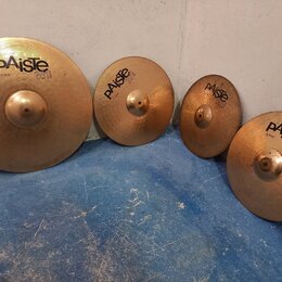 Čtyři mosazné činely značky Paiste v různých velikostech. Na činelech jsou čitelné nápisy „paiste 2002“ a konkrétní označení: „20" RIDE“, „16" CRASH“…
