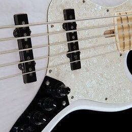 Detail bílé elektrické basy s hmatníkem ze světlého dřeva. Nástroj má bílý perleťový pickguard, dva černé snímače a černý ovládací panel s knoflíky.…