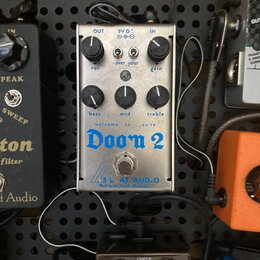 Stříbrný efekt pedál s nápisy „OUT“, „9V DC“, „IN“, „vol“, „gain“, „bass“, „mid“ a „treble“. Uprostřed je modrý nápis „welcome to ,you're Doom 2“ a v…