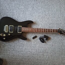 Černá elektrická kytara s tmavým hnedým hmatníkem a dvěma humbuckery. Na hlavě nástroje je bílý nápis Ibanez. Vedle kytary leží černý napájecí adapté…