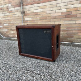 Hnědý basový reproduktor v dřevěném korpusu s černou textilní maskou. V pravém horním rohu masky je umístěn štítek s nápisem „gbr Custom Cabinets“. P…