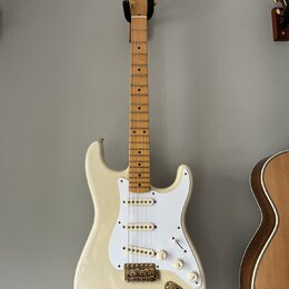 Bílá elektrická kytara s jasným dřevěným hmatníkem a bílým pickguardem. Na hlavě krku je viditelné logo Fender. Nástroj má tři snímače s bílými krytk…