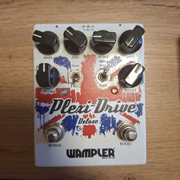 Bílý kytarový efekt s červenými a modrými grafickými prvky. Na zařízení jsou nápisy Plexi-Drive Deluxe a WAMPLER BUILT IN USA. Horní řada ovládacích…