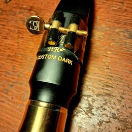 Černý saxofonový mouthpiece s mosazným prstencem u patky. Na povrchu je zlatým písmem nápis HR* CUSTOM DARK a nad ním nečitelný podpis. Mundstuk je v…
