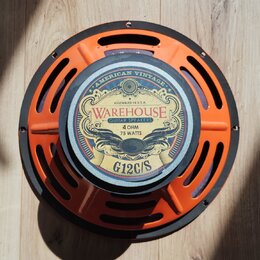 Kytarový reproduktor s oranžovým kovovým rámem a černým vnějším prstencem. Na středovém štítku je nápis WAREHOUSE GUITAR SPEAKERS, AMERICAN VINTAGE,…