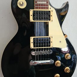 Černá elektrická kytara s dumpingovým tvarem těla a hnědým hmatníkem s trapezoidními vložkami. Nástroj má zlaté hardware, včetně dvou humbuckerů a čt…