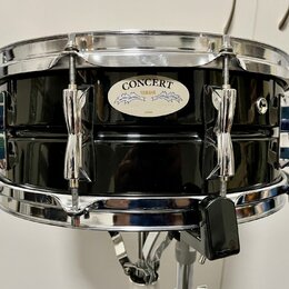 Černý buben s chromovaným obručováním a mechanismy. Na korpusu je oválný štítek s nápisy CONCERT, YAMAHA a JAPAN. Povrch nástroje je glossy, v odrazu…