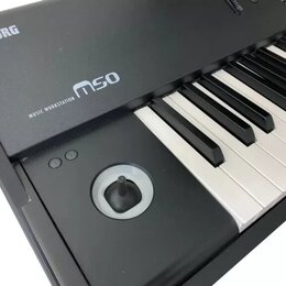 Černý klávesový nástroj s bílými a černými klávesami. Na horním panelu jsou bílé nápisy KORG a MUSIC WORKSTATION m50. V levé části je umístěn kulatý…
