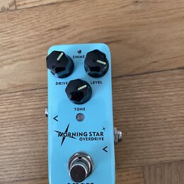 Světle modrý efekt pedál s třemi černými oválnými knoflíky. Na pedálu jsou nápisy SHINE, LEVEL, TONE a DRIVE, dále text MORNING STAR OVERDRIVE a logo…