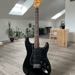 Černá elektrická kytara typu Stratocaster s černým pickguardem a hnedým hmatníkem. Na hlavě je viditelné logo „Fender Stratocaster“. Nástroj je umíst…
