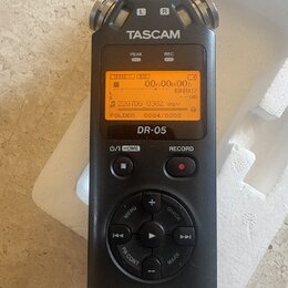 Černý digitální záznamový zařízení s nápisem TASCAM a označením modelu DR-05. Displej je zapnutý s oranžovým podsvícením a zobrazeným textem 00H00M00…