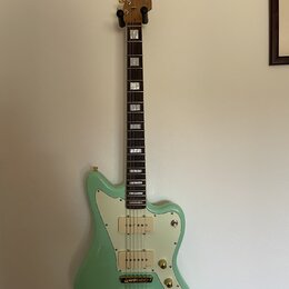 Světle zelená elektrická kytara s bílým pickguardem a zlatými potenciometry a hardwarem. Na hlavě hmatníku je viditelné logo "Epiphone". Hmatník je t…