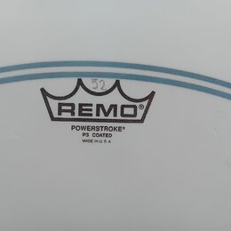Detail bílého, matného bubenového koženu s modrými vodorovnými pruhy. Uprostřed je černý nápis REMO pod symbolem koruny a text POWERSTROKE® P3 COATED…
