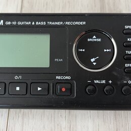 Černý digitální přístroj s nápisem TASCAM GB-10 GUITAR & BASS TRAINER / RECORDER. Zařízení má LCD displej, ovládací kolečko s nápisem BROWSE a řadu t…