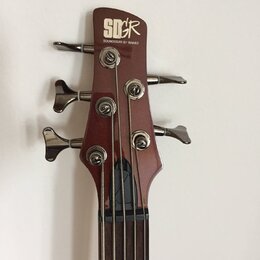 Fotografie zachycuje hlavu hnedý basové kytary s четыřmi ladicími kolíky. Na horní části je bílý nápis „SOUNDGEAR“ v stylizovaném logu a pod ním menš…