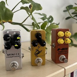 Tři efektové pedály v řadě na světlém dřevěném povrchu. První je stříbrný s nápisem MINI C4 compressor a čtyřmi černými knoflíky. Prostřední je zlatý…