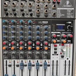 Šedý mixážní pult značky BEHRINGER s modelem XENYX X2022USB. Zařízení obsahuje několik vstupních kanálů s regulačními knoflíky, posuvníky hlasitosti…