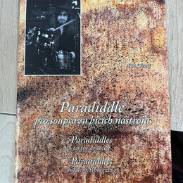 Fotografie znázorňuje obal knihy nebo sešitu s texturou připomínající rezavý kov. V levém horním rohu je černobílá fotografie bubeníka za bicími nást…