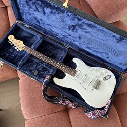 Bílá elektrická kytara typu Stratocaster v otevřeném tvrdém kufru s modrým polstrováním. Na hlavě hmatníku je viditelné logo Fender. Kytara má tři sn…