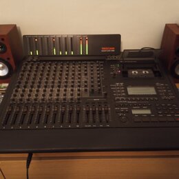 Černý digitální mixážní pult s řadou posuvníků a ovládacích knoflíků. V horní části je umístěn modul s LED indikátory a nápisem TASCAU DIGITAL STUDIO…