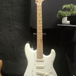 Bílá elektrická kytara typu Stratocaster s jasným dřevěným hmatníkem a hlavou. Na hlavě je viditelné logo Squier. Nástroj má tři snímače typu single-…