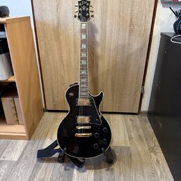 Černá elektrická kytara s gold hardwarem umístěná na stojanu. Na hlavě hmatníku je viditelné logo Epiphone. Kytara má dva humbuckery, čtyři ovládací…