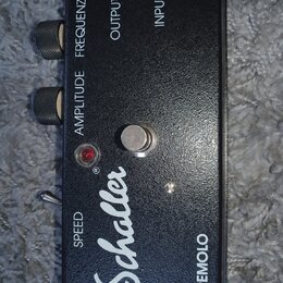 Černý efekt pedál s nápisem Schaller a TREMOLO. Na horní hraně jsou dva ovládací knoflíky s popisky AMPLITUDE a FREQUENZ, dále nápisy OUTPUT a INPUT.…