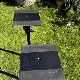 Dvě černé kovové platformy na stojanech, pravděpodobně držáky na noty nebo tablety. Povrchy jsou matně černé, přičemž spodní platforma vykazuje vidit…