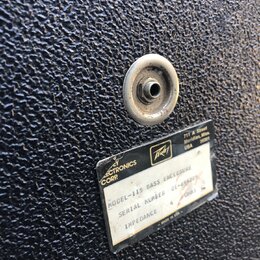 Detail černého povrchu basového komba s texturou. Na povrchu je kovový konektor a štítek s nápisy: „ELECTRONICS CORP“, logo „Peavey“ a adresa „711 A…
