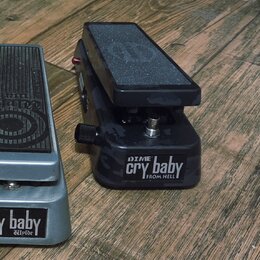 Dvě wah pedály umístěné na dřevěném povrchu. Pravý pedál má černý maskáčový vzor a nápis DIME cry baby FROM HELL. Levý pedál je stříbrný s nápisem cr…