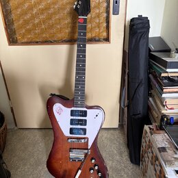 Elektrická kytara s hnědým trupem z dřeva a bílým pickguardem. Na pickguardu je červenobílý nápis "Rickenbacker". Trup vykazuje viditelné opotřebení…