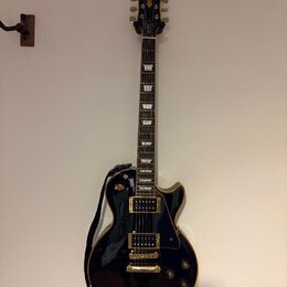 Černá elektrická kytara typu Les Paul se zlatým hardwarem a bílými blokovými vložkami na hmatníku. Na hlavě je viditelné logo Epiphone. Kytara je zav…