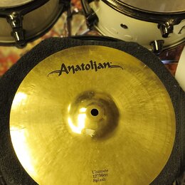 Zlatý cymbál typu splash položený na černém texturovaném podkladu. V horní části je černé logo „Anatolian“. Dole je nápis „Ultimate 12"/30cm Splash“.…
