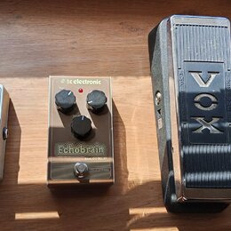 Na dřevěném povrchu leží tři kousky vybavení. Vlevo je stříbrný efektový pedál s nápisy „NEO CLONE“ a „electro-harmonix“. Uprostřed je stříbrný pedál…