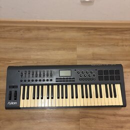 Šedý MIDI kontrolér s klávesnicí, faderem a padovými políčkami. Na levé straně je bílý nápis Axiom a v pravém horním rohu logo M-AUDIO. Povrch nástro…