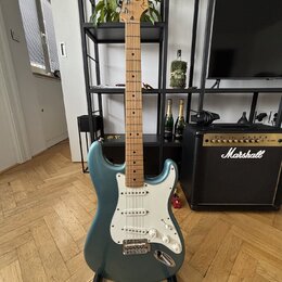Modrá elektrická kytara typu Stratocaster s bílým pickguardem a světle hnědým hmatníkem. Na hlavě je viditelné logo Fender. Kytara stojí na černém st…