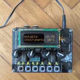 Černý elektronický modul s LCD displejem, několika otočnými knoflíky a řadou bílých tlačítek v dolní části. Na displeji je text „mutable instruments…