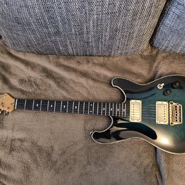 Elektrická kytara s modro-černým přechodovým lakem a krčkem z tmavého dřeva. Na hlavě je viditelné logo Ibanez a nápis RG SERIES. Nástroj má dva humb…