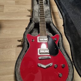 Červená elektrická kytara s černým pickguardem v otevřeném kufru s šedou vnitřní výstuhou. Na hlavě hmatníku je bílý nápis Gretsch. Kytara má dva hum…