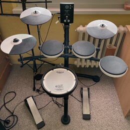 Elektronická bicí souprava značky Roland s černým rámem a bílými okraji padů. Na modulech, činelech a basovém bubnu je vidět nápis Roland; na basovém…