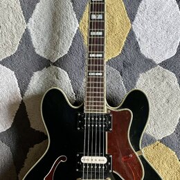 Černá polodublá elektrická kytara s bílým lemováním a hnědým hmatníkem. Na hlavě je bílý nápis Epiphone a dekorativní ornament. Tělo má dva humbucker…