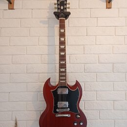 Elektrická kytara s červeným trupem v charakterickém tvaru SG a černým pickguardem. Na hlavě hmatníku je bílý nápis Gibson. Nástroj má dva humbuckery…