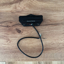 Černý humbucker s plastovým krytem a viditelnými póly. Na spodní části je bílý nápis Seymour Duncan. Z nástroje vede černý kabel s odhalenými barevný…