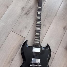 Černá elektrická kytara s dvojsekyrou v tvaru SG. Na hlavě hmatníku je bílý nápis Urville a zlatý znak. Nástroj má dva humbuckery, stříbrný most s ko…