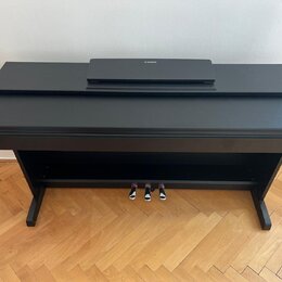 Černý digitální piano v şeklinde nábytkového kusu s uzavřenou klaviaturní víkem. Na horní desce je položen černý krycí prachový kryt s bílým logem YA…