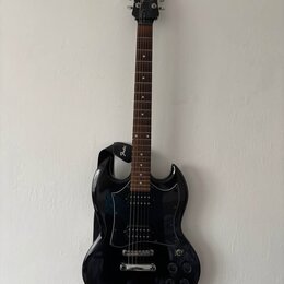 Černá elektrická kytara s tmavým hnedým hmatníkem a černým pickguardem. Na hlavě nástroje je bílý nápis Epiphone. Kytara má připevněný černý popruh s…