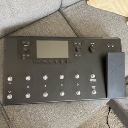 Černý kytarový procesor s displejem a ovládacími prvky. Na panelu jsou nápisy PRESET, VIEW, VOLUME, MODE HOLD TO EDIT a TAP TUNER, dále označení tlač…