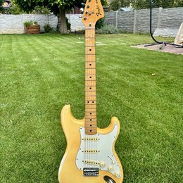 Elektrická kytara s žlutým trupem, bílým pickguardem a hnedým javorovým hmatníkem. Na hlavě je vidět logo Fender a nápis STRATOCASTER. Nástroj má tři…