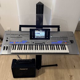 Šedý digitální klavír s logem YAMAHA na horní části a označením Genos na pravé straně. Nástroj je umístěn na černém stojanu a nad ním je průhledlá hu…