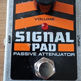 Kytarový pedál v stříbrném kovovém provedení s černým a oranžovým panelem. Na panelu je nápis SIGNAL PAD PASSIVE ATTENUATOR, označení VOLUME u ovláda…